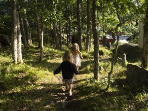 To små jenter løper på en sti i en skog. Trærne lager skygger over stien, som ellers ligger i sollys.