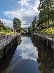 Bilde av et slusekammer fylt med vann. På den ene siden av slusa ligger en gul bygning. På den andre siden av slusa ligger en liten rød bod. Himmelen speiler seg i vannet i slusekammeret.