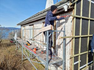 Et bilde av en mann som står på et stillas og holder et brekkjern mot kledningen på bygningen.