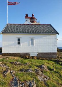 Bilde av en hvit bygning med grått skifertak. Foran bygningen er det grønt gress, og bak bygningen er himmelen blå. Man kan også se en fyrlykt i bakgrunnen. Fra en flaggstang vaier en vimpel i vinden.